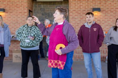 A woman wearing a maroon Virginia Tech apron gestures while speaking to a small group gathered under a brick pavilion